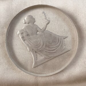May include: A clear glass cameo with a raised design of a woman sitting in a chair. The woman is wearing a long dress and holding a flower.