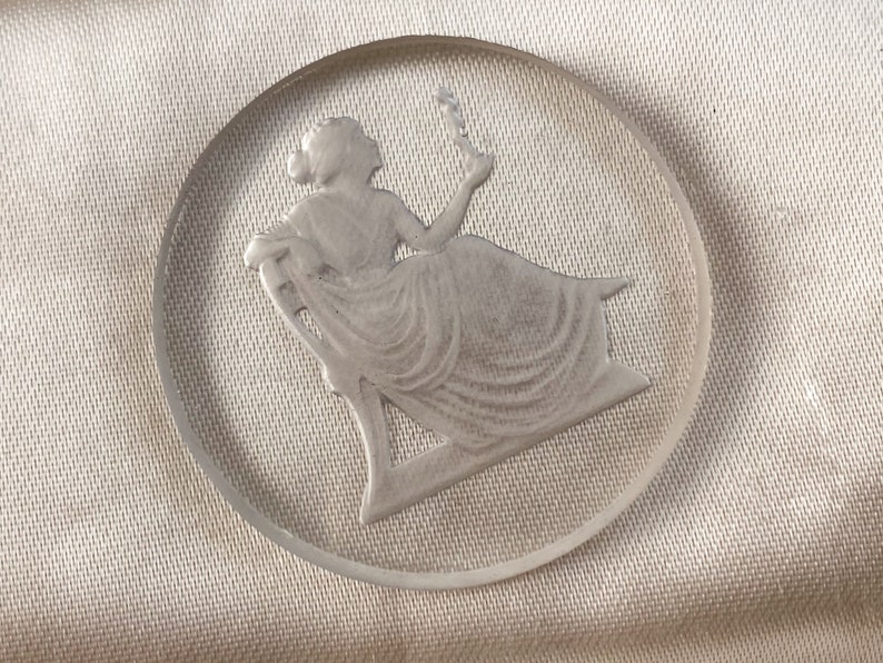 May include: A clear glass cameo with a raised design of a woman sitting in a chair. The woman is wearing a long dress and holding a flower.