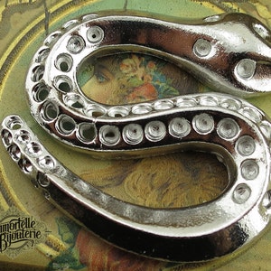 May include: A silver-colored snake-shaped pendant with a textured surface and a row of circular cutouts along the body.
