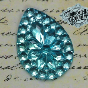 May include: A light blue glass teardrop-shaped cabochon with a raised, starburst design. The cabochon has a shiny, iridescent finish.