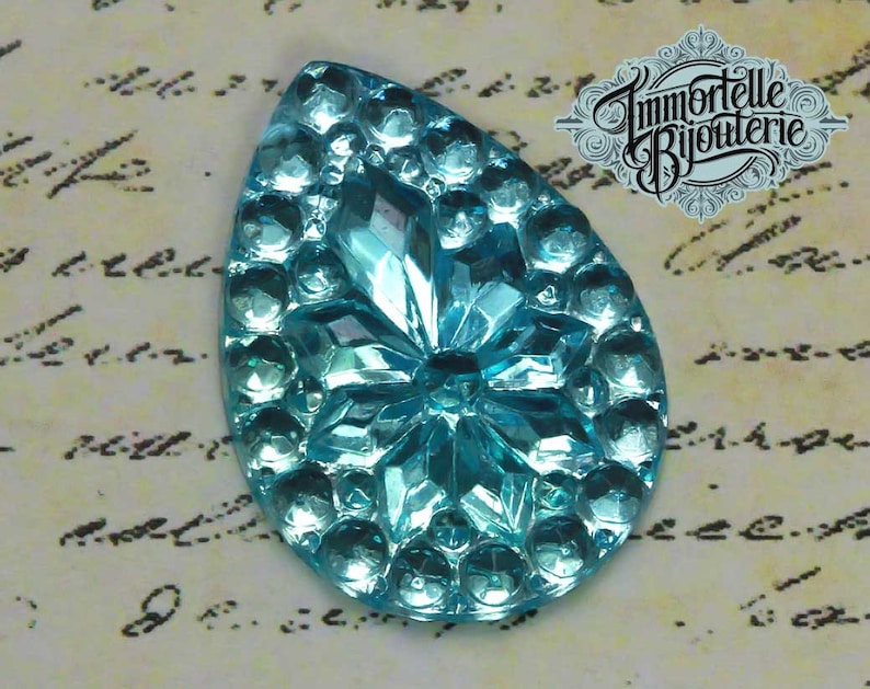 May include: A light blue glass teardrop-shaped cabochon with a raised, starburst design. The cabochon has a shiny, iridescent finish.