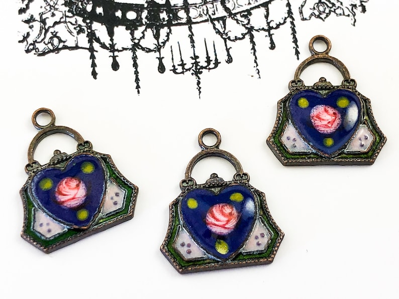 May include: Three antique-style metal charms shaped like purses with a heart-shaped front. The hearts are painted blue with a pink rose and yellow dots. The purses have green and white accents.