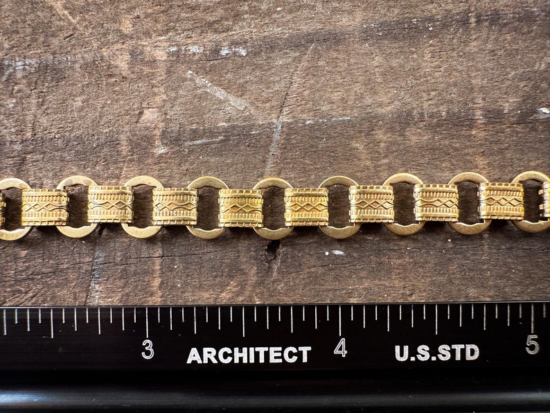 K&ouml;nnte beinhalten: Ein goldenes Kettenarmband mit geometrischem Muster. Die Kette besteht aus miteinander verbundenen Kreisen mit einem dekorativen Design.