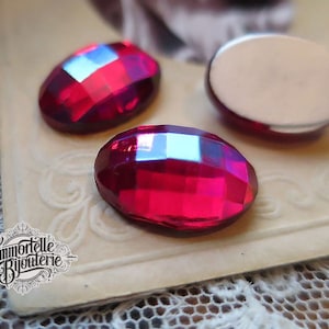 May include: Close-up of three faceted, oval-shaped, ruby-red glass cabochons. The cabochons have a flat, silver-colored backing. The image includes the logo "Immortelle Bijouterie" in a vintage style.