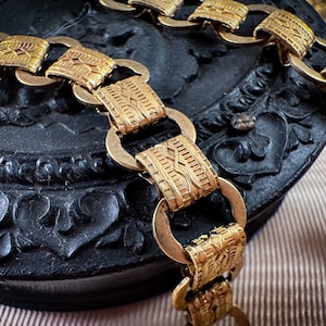 K&ouml;nnte beinhalten: Eine goldene Kettenkette mit einem detaillierten, komplizierten Design. Die Kette besteht aus miteinander verbundenen ovalen Formen mit einer strukturierten, gemusterten Oberfl&auml;che.