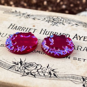 May include: Two round, iridescent glass buttons with a raised, textured edge. The buttons are a deep red colour with a blue iridescent sheen. The buttons are sitting on a vintage box with the text "Harriet Hubbard Ayer" printed on it.