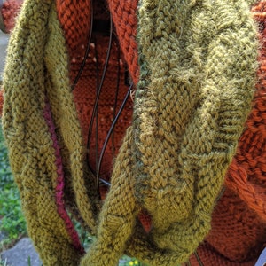 May include: A green and orange knitted scarf with a textured pattern. The scarf is folded and draped over a metal rack.