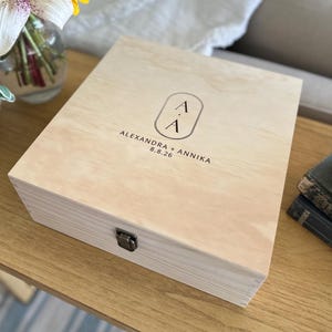 May include: A square light-colored wooden keepsake box with a metal latch on the front. The lid features a black printed design with a vertical oval containing the letters A and A, followed by the text ALEXANDRA + ANNIKA and the date 8.8.26.