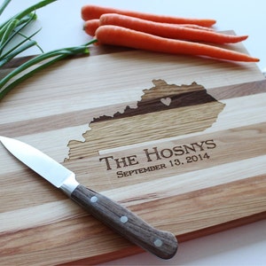 May include: A wooden cutting board with a personalized inscription that reads "The Hosnys September 13, 2014". The board is made of multiple wood types and features a heart-shaped outline of the state of Kentucky.