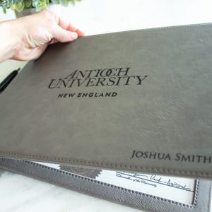 May include: Gray leather folder with the text "Antioch University New England" and "Joshua Smith" embossed on the front.