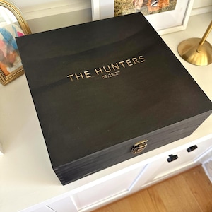 May include: A square black wooden box with a hinged lid and a gold metal latch. The top of the lid features gold lettering that reads THE HUNTERS followed by the date 05.29.27. The box has visible wood grain and a dark matte finish.