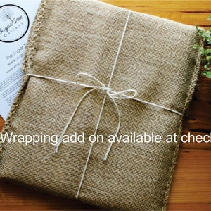 May include: A brown burlap gift bag with white twine tied in a bow. The bag is tied with a white ribbon and has a white tag with the text "SugarTree Gallery" on it. The text "Gift Wrapping add on available at checkout" is written below the bag.