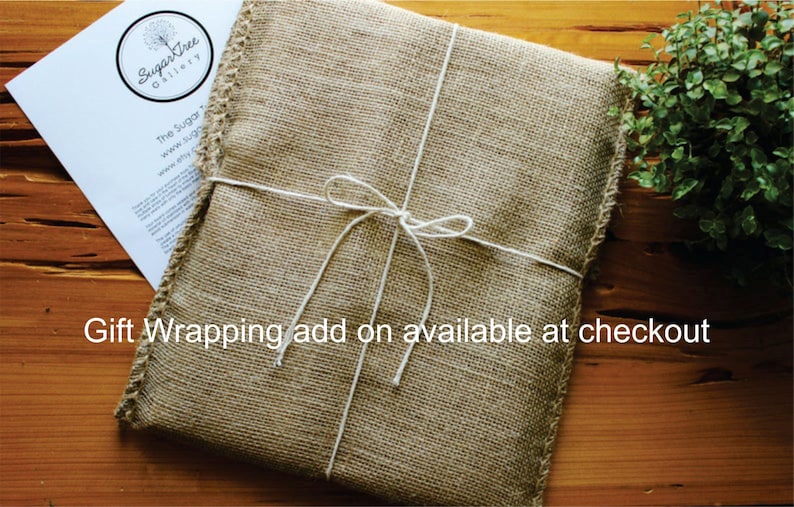 May include: A brown burlap gift bag with white twine tied in a bow. The bag is tied with a white ribbon and has a white tag with the text "SugarTree Gallery" on it. The text "Gift Wrapping add on available at checkout" is written below the bag.