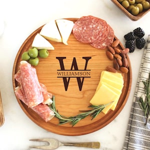 May include: A round wooden serving board with a personalized monogram of the letter "W" and the name "Williamson" engraved on it. The board is filled with various snacks, including cheese, salami, olives, and almonds.