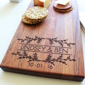 May include: A personalized wooden serving board with the names Lindsey & Ben and the date 10-01-16 engraved on it. The board is decorated with a floral design.