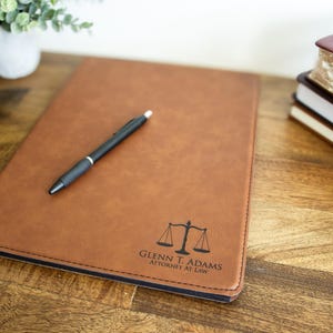 May include: A brown leather portfolio with a black pen on top. The portfolio has a scale of justice and the text "Glenn T. Adams Attorney at Law" embossed on the front.