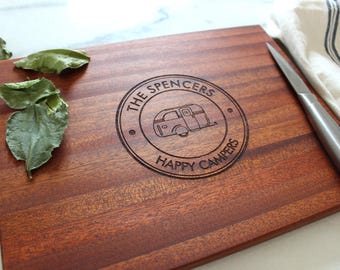 May include: A rectangular wooden cutting board with a dark reddish-brown grain. The center features a circular laser-engraved design of a vintage camper trailer with the text THE SPENCERS and HAPPY CAMPERS. A silver knife rests on the right side.