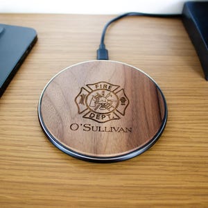 May include: A round wooden wireless phone charger with a black trim. The charger features a fire department emblem and the name "O'Sullivan" engraved on the wood. A black smartphone and charging cable are also visible.