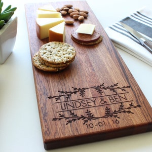 May include: A personalized wooden serving board with the names Lindsey & Ben and the date 10-01-16 engraved on it. The board is brown and has a natural wood grain. There are crackers, cheese, and almonds on the board.