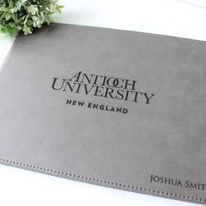 May include: Gray leather portfolio with the text "Antioch University New England" and "Joshua Smith" embossed in black.
