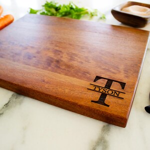 May include: A rectangular wooden cutting board with the name "TYSON" engraved. The board is a warm brown color and has a knife with a black handle next to it. Carrots and herbs are also visible.