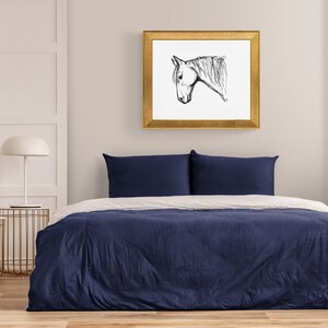 May include: A navy blue duvet cover on a bed with two navy blue pillows. A framed black and white sketch of a horse's head hangs on the wall above the bed. A white lamp and a vase of purple flowers are on the nightstand.