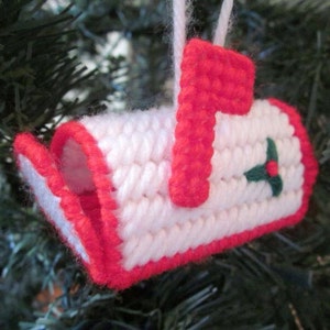 Puede incluir: Adorno de buzón de correo de crochet blanco y rojo con una rama de acebo verde. El adorno está colgado de un árbol de Navidad.