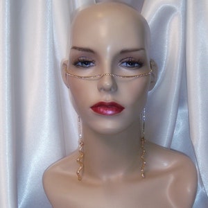 May include: A mannequin wearing a gold chain nose ring and long, dangling gold earrings with rhinestones. The mannequin is posed against a white satin background.