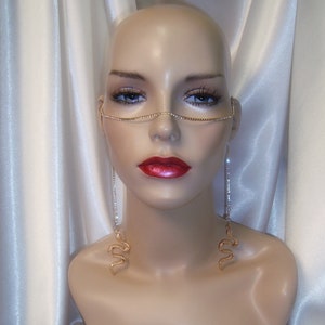 May include: A mannequin head displays a gold-toned face chain and dangling earrings. The face chain is adorned with small, sparkling stones. The earrings feature a snake design, with a long, dangling chain of stones.