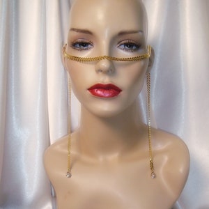 May include: A gold-colored face chain with a chain design across the bridge of the nose and dangling chains with crystal accents. The face chain is displayed on a mannequin head with red lipstick and a white satin background.