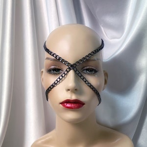 May include: A mannequin head with a black chain face mask. The mask is in an X-shape across the eyes and face. The mannequin has red lipstick and is set against a white satin background.