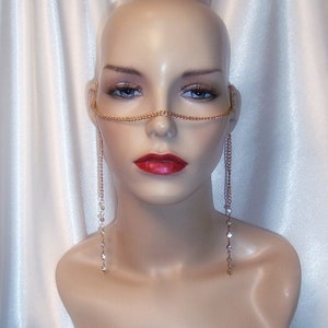 May include: A gold-toned face chain with dangling crystal accents. The jewelry piece is designed to be worn across the forehead and around the ears, with delicate chains of crystals hanging down. The mannequin has red lipstick and is set against a white satin background.
