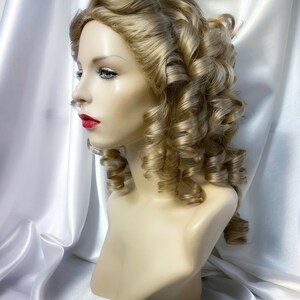 May include: A blonde wig with large curls styled in a vintage updo. The wig is on a mannequin head and is displayed against a white satin background.