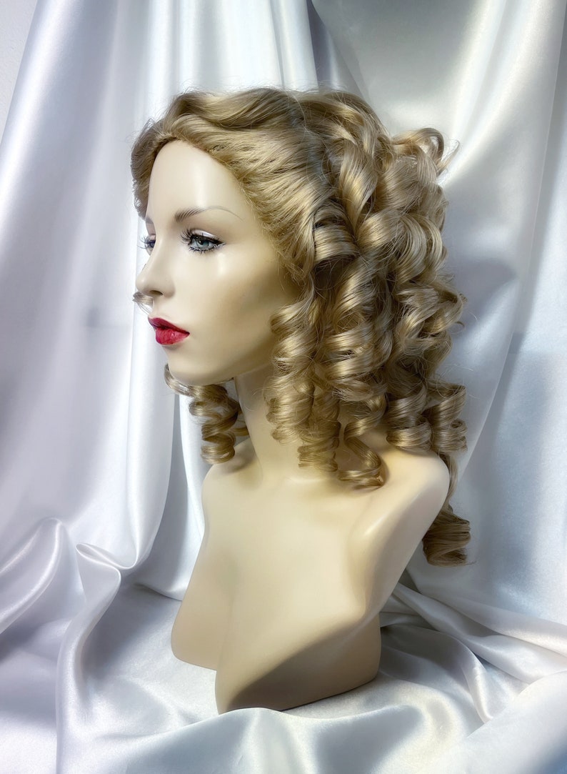 May include: A blonde wig with large curls styled in a vintage updo. The wig is on a mannequin head and is displayed against a white satin background.