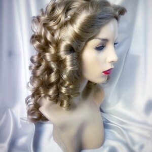 May include: A blonde wig with large curls styled in a vintage updo. The wig is on a mannequin head and has a few small white pearl accents.