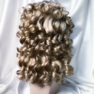 May include: A blonde wig with large, loose curls styled in a classic updo. The wig is on a mannequin head and is set against a white background.