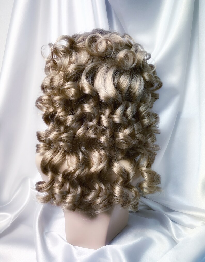 May include: A blonde wig with large, loose curls styled in a classic updo. The wig is on a mannequin head and is set against a white background.