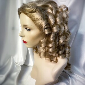 May include: A blonde wig with large, loose curls styled in a classic updo. The wig is on a mannequin head and is set against a white background.