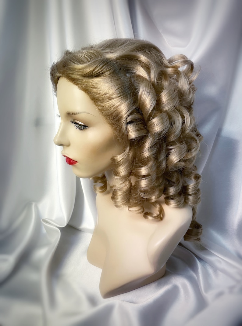 May include: A blonde wig with large, loose curls styled in a classic updo. The wig is on a mannequin head and is set against a white background.