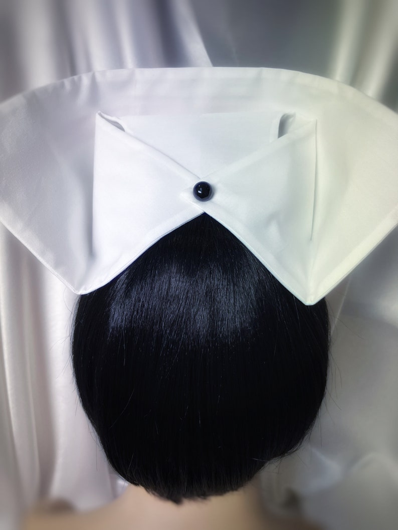 May include: White nurse's cap with a black button on the front. The cap is folded in a triangular shape and is worn over a black ponytail.