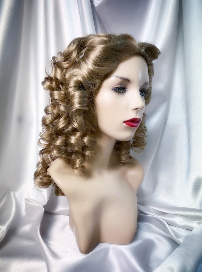 May include: A blonde wig with large, loose curls styled in a vintage updo. The wig is on a mannequin head and is set against a white satin background.