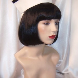May include: A white nurse's cap with a black band around the crown. The cap is on a mannequin with black bobbed hair and red lipstick.