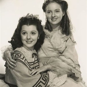 May include: Two young women in vintage dresses, one with a floral headband and the other with a floral crown, pose together in a studio portrait. The woman in the front is wearing a dress with a geometric pattern.