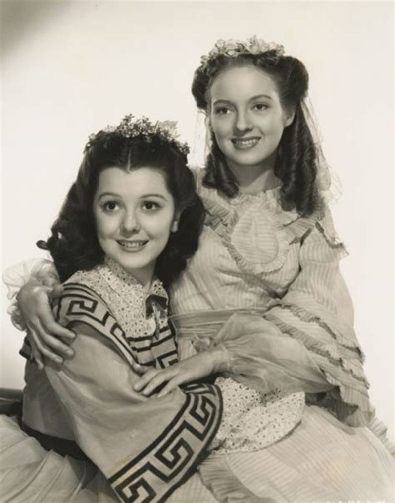 May include: Two young women in vintage dresses, one with a floral headband and the other with a floral crown, pose together in a studio portrait. The woman in the front is wearing a dress with a geometric pattern.