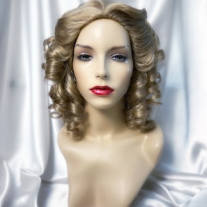 May include: A blonde wig with loose curls styled in a vintage updo. The wig is on a mannequin head and has a natural-looking finish.