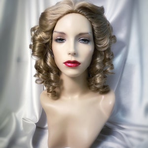 May include: A blonde wig with loose curls styled in a vintage updo. The wig is on a mannequin head and has a natural-looking finish.