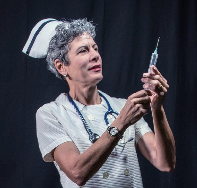 May include: A woman wearing a white nurse's uniform and a white cap with a blue stripe holds a syringe with a needle. She is wearing a watch on her left wrist and a stethoscope around her neck.