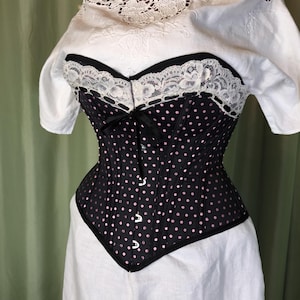Può includere: Un corsetto nero a pois rosa, rifinito con pizzo nero e bianco. Il corsetto ha una scollatura a cuore ed è impreziosito da un fiocco nero. È abbinato a una camicetta bianca.