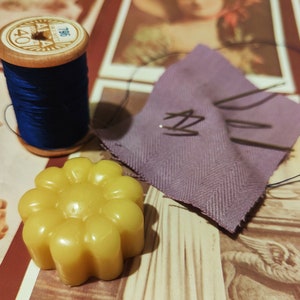Puede incluir: Una bobina de hilo azul con una etiqueta blanca que dice "40" y "756", una tela morada con algunos alfileres y una vela amarilla con forma de flor.