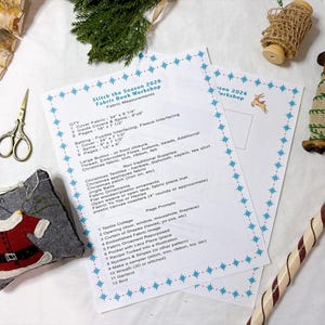 Op de afbeelding: Twee gedrukte instructiebladen getiteld Stitch the Season 2026 Fabric Book Workshop met blauwe sterrenranden, vergezeld door een gouden schaar, een grijs vilt speldenkussen, een houten klosje met groene afwerking en een zuurstok.